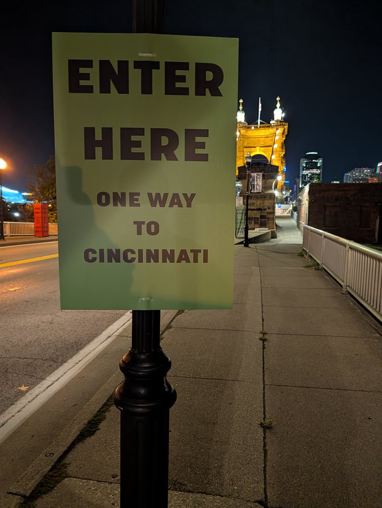 Lamp post sign reading Enter Here One Way to Cincinnati with the illuminated Roebling Bridge tower in the background